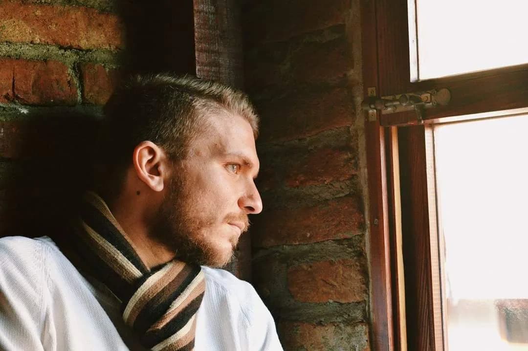 Teoman Maksut in profile, looking out a window against an exposed brick wall.
